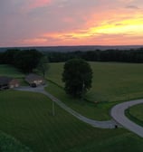 Aerial view of a country home surrounded by open fields, framed by a beautiful sunset in the sky