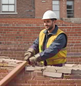 A person is standing among stacks of large, rectangular stone slabs arranged in an outdoor setting. The individual is wearing a white shirt and black pants, with one hand positioned under their chin as they appear to be in thought. In the background, there are power lines and a sparse amount of greenery.