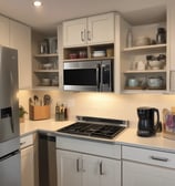 A retro-style kitchen setup featuring a pastel blue microwave and a cream-colored toaster stacked on top. Next to them is a matching pastel blue kettle. The appliances are placed on a wooden countertop against a white tiled wall.