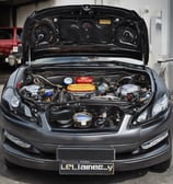 A classic red car is parked in front of an auto repair shop, flanked by two modern vehicles. The auto shop has a sign in both English and another language, featuring a logo similar to BMW. The area appears slightly wet, indicating recent rain.