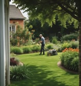 A residential house with a red gate and lush foliage in the foreground. A person is sweeping or cleaning leaves off the pavement. Another person can be seen sitting near the gate, possibly resting. The scene includes vibrant pink flowers partially obscuring the view, and the area is shaded by trees.