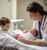A woman in a white coat is assisting a young child sitting on another woman's lap during an eye examination. The child is positioned in front of a medical device for eye testing, with the woman holding her in place. The setting appears to be an eye clinic room.