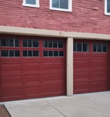 A shirtless man is painting the exterior wall of a white building using a paint roller on a long pole. The building has a wooden window on the left and another entrance with a red railing on the right. There are some cardboard pieces on the floor next to the wall, and a few other people are walking in the background.