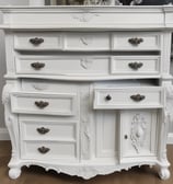 A person wearing a pink shirt and plaid shorts is balancing on the edge of an open window while painting the upper part of the frame. The window has a green frame with multiple small circular panes at the top. There is a paint can and brush being used for painting. The curtains inside the room are drawn, and the overall wall color is a peach or light orange.