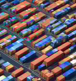 A large cargo ship is sailing across a calm body of water under a mostly cloudy sky. The vessel is fully loaded with colorful shipping containers stacked on its deck. In the background, a small sailboat is visible on the left side of the image.