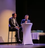 Deux personnes assises sur des tabourets sur scène qui discutent avec un micro