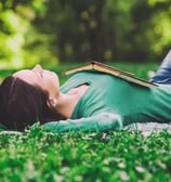 Femme allongée dans l'herbe, jambes repliées, pieds sur le sol et un livre ouvert sur sa poitrine