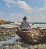 Femme assise sur un rocher face à la mer, mains écartées, doigts joints en posture de méditation