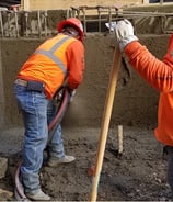Concrete workers wearing orange safety vests and hard hats, one holds a hose, the other a shovel.