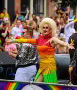 A drag queen in a rainbow dress with large blonde wig holds her arms out in greeting