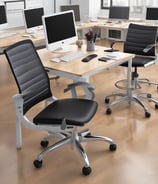 A modern office setup with a sleek ergonomic chair in the foreground. The chair features a complex support structure and black cushioning. Behind it, there is a computer desk with a large monitor displaying a bright screen and a microphone on an adjustable arm. A keyboard and phone can also be seen on the desk. Some greenery is present in the background, adding a natural element to the workspace.