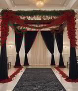A canopy with ornate drapes and floral arrangements at the entrance. The interior features carpeted flooring with intricate patterns, leading to an event space where people are gathered. The canopy is adorned with lush greenery and pink flowers, creating an elegant and welcoming atmosphere.