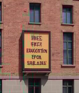 A child is holding a cardboard sign with the message 'Fund Childcare Not Wars' written on it, featuring colorful scribbles and drawings. The background shows a city street with blurred people and lights, suggesting a protest or demonstration setting.