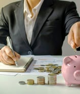 a person in a suit jacket is depositing money from one hand & holding a pen and writing on a notepad