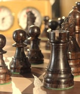 Closeup view of a chess board with black pieces and a chess clock in the background.