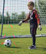 A young person is standing on a soccer field with one foot resting on a yellow soccer ball. They are wearing a gray soccer jersey and black shorts with knee-high socks. The background features a net and a wooden wall with a blue sky overhead.