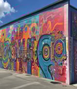 A modern gallery space featuring various art pieces on display. A large video screen shows a close-up image of an older man with a worn expression, wearing a hat and a blue shirt. Two people are in the gallery, one person standing and looking at their phone while another is walking with hands in pockets. Photographs are hung on the wall in the background, depicting outdoor scenes.