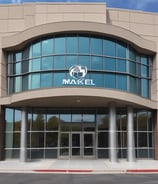 A sign with the text 'MIKAJA' and 'Agencia de envios a Cuba' attached to a brick wall. The sign features a stylized logo with a red and white box, a blue arrow, and a white star on a red background. The brick wall has several patches, some exposed wires, and a partially visible arched window.