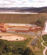 Vista aérea de uma barragem com vertedouro em funcionamento, cercada por áreas verdes e montanhas ao fundo