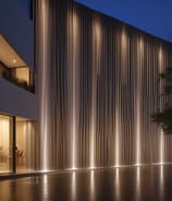 Three outdoor spotlights are mounted on a metal bracket against a building facade. The background is blurred with pastel-colored walls and windows, providing a soft contrast to the industrial lighting fixtures.