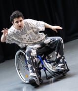 A mixed race manual wheelchair user, leaning to the side, arms spread out, and one foot in front