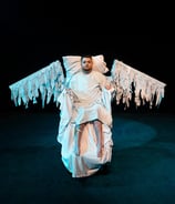 An electric wheelchair user in a hospital gown, with swooping wings made of the same fabric