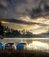 Tiga perahu bersandar di tepi Danau Situ Cileunca saat matahari terbit, dengan kabut tipis dan pantulan langit di permukaan a