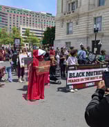 Front of the marching line. People are holding a banner and a woman is dressed like a Handmaid. 