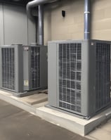 Three air conditioning units mounted on the exterior wall of a building, with a closed vent in the center. The wall is a warm, peach color and the units have a beige finish. A black cable runs vertically along the left side of the image.