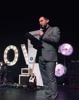 Alex hosting an awards function in a grey suit holding a clipboard