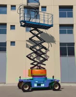 A large boom lift truck is parked on a city street next to a tall building. The truck has its arm extended with a platform at the end. Several orange traffic cones surround the vehicle, indicating an area of work or caution. Two workers appear to be standing on the platform of the boom lift, engaged in some sort of maintenance or construction activity.