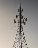 A large telecommunications tower is equipped with numerous antennas and satellite dishes. The structure is complex, with cables and metal components extending outward, set against a background of partly cloudy skies.