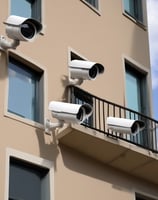 A red brick building facade featuring a distinctive white cornice at the top. A surveillance camera is mounted above a large wooden door, which is flanked by a vintage-style exterior light fixture. An ornate railing runs horizontally across the lower part of the image.