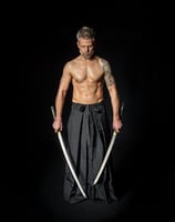 Ein muskulöser, tätowierter Mann mittleren Alters steht mit zwei Katanas auf einem schwarzen Hintergrund. Er trägt einen trad