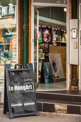 A sandwich board on the sidewalk, pointing to the entry of an art gallery with paintings inside