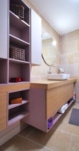 Une salle d'eau dont les murs sont carrelés de travertin, les meubles en bois, le miroir rond.