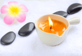 a massage candle in a cup container over a white cloth table with mindfulness stone and a flower