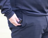 Golfer in black athletic wear placing a black and orange golf rangefinder into the right pocket of their trousers.