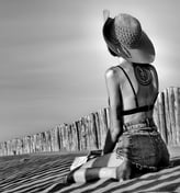 Photo artistique noire et blanc d'une jeune femme en mini short a genoux sur la plage avec un livre et un grand chapeau