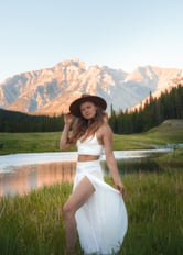 a woman in a white dress and hat standing in the grass with a mountain in the back in Banff