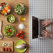 Person typing on laptop at other end of table that displays many dishes/ingredients
