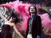 A model dressed as the Joker poses while a Harley Quinn spreads purple smoke in the background