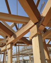 A construction scene with workers building a multi-story wooden structure. Two people are on the roof and another is on a lift, all actively engaged in the building process. The unfinished structure has exposed wooden frames and large window openings.