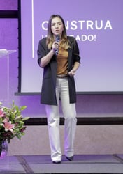 uma mulher em pé encima de um palco, em frente a um projetor de imagens com um microfone