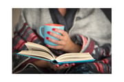 A woman wrapped in a blanket reads a book and holds a mug