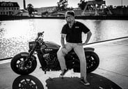 Ein Schwarz-Weiß-Foto eines Mannes, der auf einem Motorrad sitzt und über einen Gewässer blickt. Der Mann trägt eine Sonnenbr