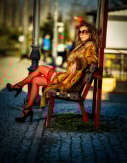 Eine Frau mit lockigen Haaren, Sonnenbrille, Pelzmantel und roten Strümpfen sitzt auf einer roten Bank im Freien.