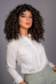 Porträt einer Frau mit Locken, blauen Augen und weißem Hemd, die über die Schulter blickt.