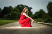 Eine junge Frau im roten Abendkleid hockt auf einem Weg mit Bäumen im Hintergrund.