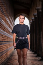 Eine selbstbewusste Frau in schwarzem T-Shirt, Fischernetzstrumpfhose und Gürtel, die in einem gemauerten Gang steht.